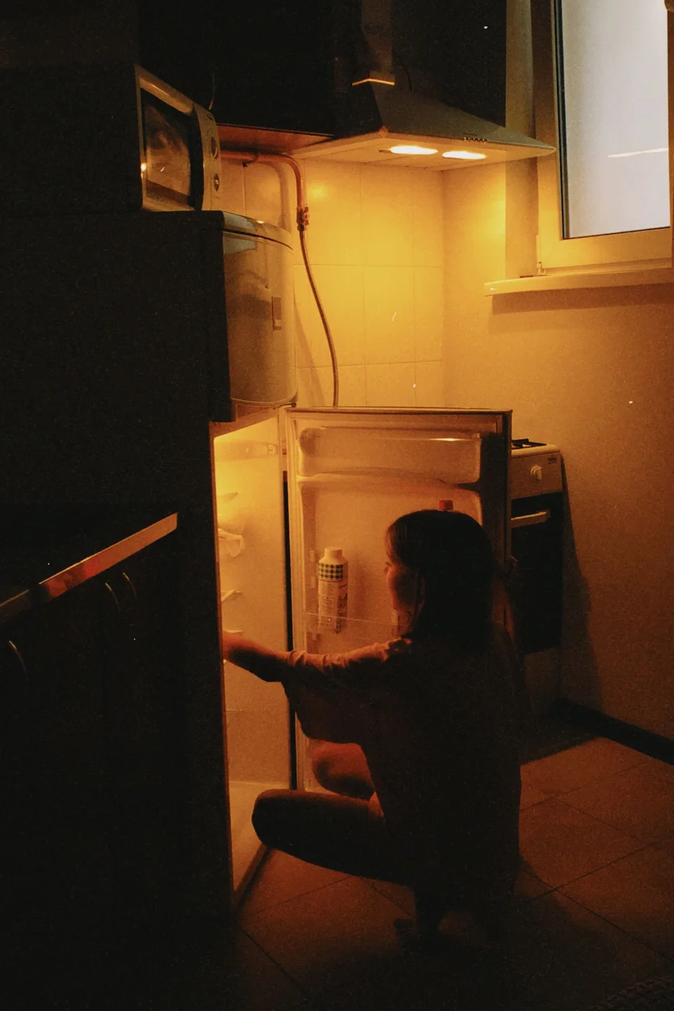 Silhouette accroupie devant un réfrigérateur ouvert la nuit, évocation du rapport à l'alimentation